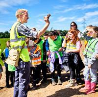 Man wearing a hi-viz vest holding a small object in his hand, standing in front of a group of children also in hi-viz vests. 